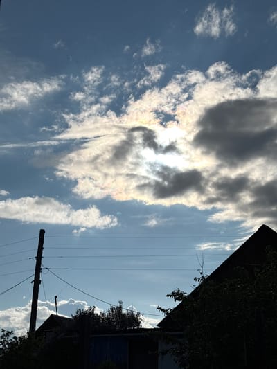 The image captures a sky filled with dramatic clouds, some tinged with iridescent colors suggesting a rainbow effect or unusual atmospheric conditions. The sun is partially obscured by thick clouds, creating a strong contrast between light and shadow. In the foreground, silhouetted against the sky, are the rooftops of buildings and the tops of trees. A utility pole with wires stretches vertically on the left side of the frame. The scene appears to be an outdoor, possibly rural or suburban, environment. The specific location in Bratsk, Russia, is not discernible from the visual elements present.