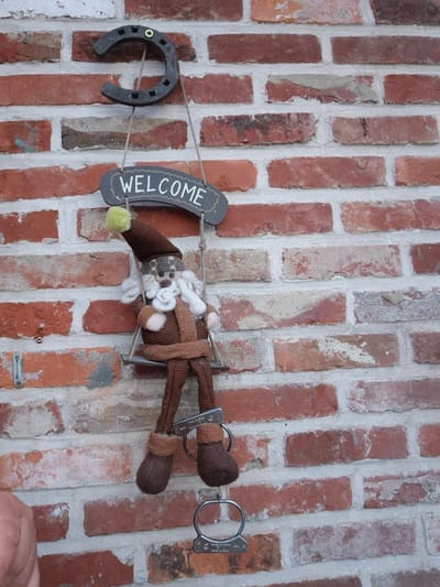 An outdoor shot focuses on a whimsical decoration attached to a red brick wall. A horseshoe is mounted at the top, with two ropes descending from it. These ropes suspend a small wooden swing on which a plush gnome is seated. The gnome is dressed in a brown, textured outfit with a pointed hat adorned with a yellow pompom. Below the gnome, a metal chain is visible, attached to its feet. The chain appears to have a small circular emblem or tag on it, as does another similar chain hanging lower. The overall context suggests a decorative item meant to welcome visitors, possibly outside a home or establishment in Aalst, Belgium, though the exact location within the city cannot be determined from the image alone.