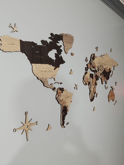 A wooden world map decorates a light gray wall. North America is shown in two shades of brown, with "Alaska," "Canada," and "Estados Unidos" etched into the wood. South America is to the lower right, also brown. Europe and Asia are on the right side. Small wooden airplanes are placed near North America and between South America and Europe. A compass rose sits to the lower left of South America.