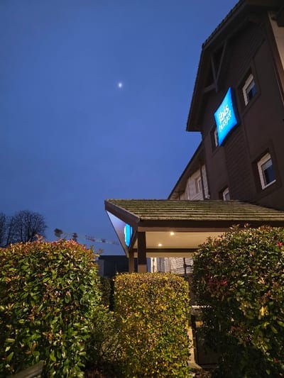 A building with a blue "ibis Styles" sign stands under a blue sky at dusk. The building has brown siding and a pitched roof. To the right, bushes with green and yellow foliage obstruct part of the building. A pale light source is visible in the sky, possibly a moon or star. A crane is visible in the distance.
