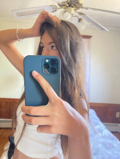 A young woman with long, brown hair takes a selfie in a bedroom in Oak Bluffs, United States of America. She is holding a dark blue smartphone in her right hand, which is raised to capture the image in a mirror. Her left arm is bent, with her hand resting on her head, partially obscuring her hair. She is wearing a white top with a black sash. The background shows a ceiling fan, wood-paneled walls, and a bed with a patterned duvet. The lighting suggests an indoor, daytime setting.