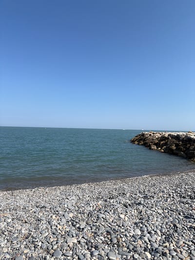 The image depicts a tranquil seaside scene on a clear, sunny day. The foreground is dominated by a vast expanse of smooth, grey pebbles that form the beach. The pebbles vary in size and shape, creating a textured surface that stretches towards the gentle waves of the sea. The water is a tranquil blue-green, with subtle ripples indicating a light breeze.
In the middle ground, a breakwater constructed from large, dark rocks juts out into the sea, creating a protective barrier. The breakwater is covered in various shades of grey and brown rocks, some of which appear wet from the encroaching waves.
Further out in the distance, the horizon is marked by a faint line where the sea meets the sky. Several small white boats or buoys are visible on the water's surface, adding a touch of distant activity to the otherwise serene landscape. The sky above is a vast, uninterrupted expanse of clear blue, devoid of any clouds.
This scene is characteristic of the coastal areas in Cagnes-sur-Mer, France, known for its pebble beaches and the protective breakwaters found along its shoreline.
