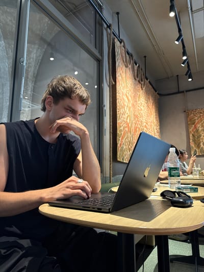 A man sits at a light-colored, round wooden table in what appears to be a café or restaurant. He is fair-skinned with short, light-brown hair, wearing a black sleeveless shirt. He is typing on a partially open laptop, and his left hand is near his face. On the table, there is a bottle of water, sunglasses, and a mouse. Behind him, a glass wall shows the outside environment. Above, track lighting is visible, along with a decorative fabric hanging from the ceiling. The café appears to be located in the Hongkou District of China.