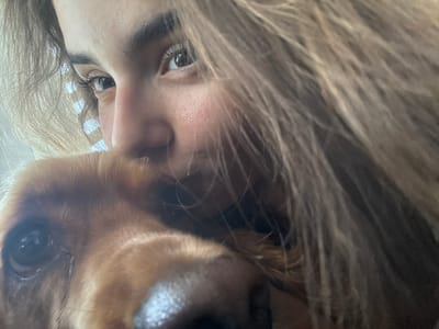 A close-up, low-angle selfie shows a young person's face partially obscured by their long, light brown hair and the head of a golden-brown dog. The person's eye, left cheek, and eyebrow are visible. The dog's snout and one eye are prominent in the foreground. The background consists of vertical blinds, suggesting an indoor setting. The image was taken in or around Dibich, Bulgaria.