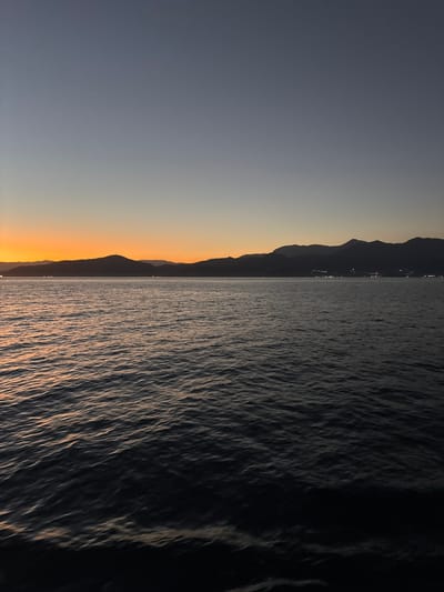 The image depicts a serene coastal landscape at dusk. The sky transitions from a deep blue at the top to a warm orange glow near the horizon, suggesting the last moments of sunset. Silhouetted against the fading light are a range of mountains, their peaks softly defined. Along the base of the mountains, scattered lights indicate a town or settlement nestled by the coast.

The foreground is dominated by a body of water, likely the sea, with gentle ripples reflecting the sky's colors. The water appears dark and calm, with the light catching the crests of small waves. The overall impression is one of tranquility and natural beauty as day turns into night.

The location is the city of Angra dos Reis, Brazil, known for its stunning bay dotted with islands and its mountainous coastline.