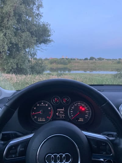 This image is taken from inside a car, looking out the windshield and at the dashboard. The subjects are the steering wheel, dashboard instruments, and the scenery outside. The dashboard displays a speedometer, tachometer, fuel gauge, and other warning lights. The speedometer indicates a speed of approximately 0 km/h, and the tachometer shows the engine is at idle. The odometer shows 229406 km, and the trip meter shows 395.0 km. A digital display shows the time as 21:14 and the date as 22.06.2026. The scenery outside the car is a rural landscape with a body of water, grassy fields, and trees. The sky is clear and blue, suggesting it is daytime, though the time of 21:14 indicates it is late evening, possibly with the sun low on the horizon or just set. The overall impression is of a vehicle stationary in a peaceful, natural setting. The Audi logo is visible on the steering wheel. The steering wheel has buttons for mode selection and other controls. The location is identified as Coșnița, Moldova. There are no people visible in the image. The scene appears calm and uneventful.
