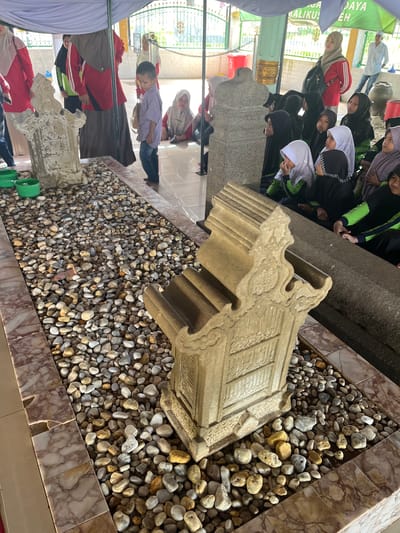 The image depicts a scene at the Sunan Ampel Mosque complex in Beuringen, Indonesia. In the foreground, a sandstone gravestone, intricately carved with Arabic calligraphy, rests on a bed of small, multi-colored pebbles. To the left of the gravestone are three green bowls, also filled with pebbles. Behind the gravestone, a long, dark, stone sarcophagus is visible. A group of people, mostly women and children wearing hijabs, are gathered in the background, some sitting and observing the gravestones. A young boy in a plaid shirt stands between the gravestone and the group of onlookers. In the far background, a man in a white shirt and jeans stands near a large clay pot. The overall environment appears to be an outdoor, covered area, possibly a cemetery or a sacred site, with patterned marble tiling on the floor and a white canopy overhead. The lighting suggests it is daytime, with some areas in shadow.