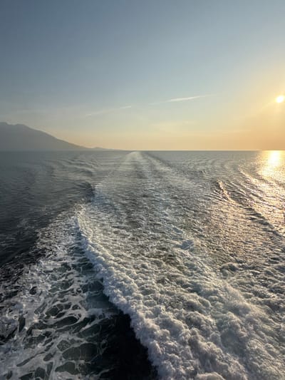 This image depicts an expansive view of open water, consistent with the sea near Kato Karioutes, Greece, under conditions indicating either sunrise or sunset. The most prominent feature is a broad, V-shaped trail of aerated, white foam constituting the wake of an unseen vessel, originating in the immediate foreground and extending directly towards the distant horizon. The water immediately surrounding the wake appears dark blue-green with gentle undulations. To the far right of the frame, a bright, circular golden sun is positioned low on the horizon, casting a shimmering, reflective path across the water's surface towards the viewer. In the upper left quadrant, a series of hazy, dark mountainous landmasses are visible along the distant horizon. The sky transitions from a pale blue at the top of the frame to a warm, yellowish-orange tint near the horizon, particularly evident on the right side. No individuals or other discernible vessels are present in the scene.