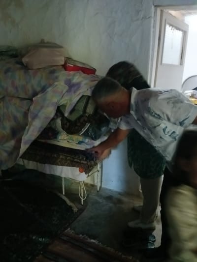 The image depicts an indoor scene, likely a bedroom or storage area, with the presence of several individuals. A man in a white patterned shirt is bent over a stack of items, which appear to be bedding or blankets covered with patterned fabric. He is reaching towards the lower portion of the stack. Behind him, another person, partially obscured, is wearing a dark, patterned garment. The room itself has rough, textured walls, and a bed frame is visible with items placed on and around it. Carpeting or rugs are present on the floor. The overall impression is of a cluttered domestic space. The metadata does not provide information about the specific location within Bahşılı Belediyesi, Turkey.