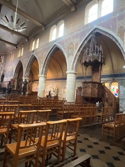 The interior of a church in Honfleur, France, is depicted. The image shows rows of empty wooden pews and a large wooden pulpit. Arched windows with stained glass are visible in the background, as is a chandelier. The walls are adorned with paintings and decorative elements. The floor is tiled in a diamond pattern. The overall impression is one of historical significance and religious ambiance.