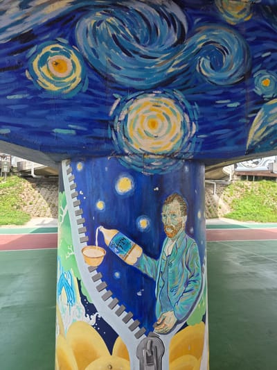 This image shows a mural painted on a pillar in Anseong-si, South Korea. The mural depicts a stylized version of Vincent van Gogh's "The Starry Night" painting on the upper portion, with swirling blue and yellow brushstrokes. Below the starry sky, the mural transitions to a portrait of Vincent van Gogh himself, looking to the left. He is wearing a blue jacket and is depicted pouring a golden liquid from a bottle into a yellow bowl. The bottle has Korean text on it, which appears to be "광해". The pillar is located outdoors, next to a sports court with green and red surfaces. Behind the pillar, a grassy slope with some concrete structures and a bridge is visible. The overall impression is a vibrant and artistic mural integrated into a public space.