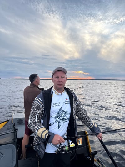 Two men are in a boat on a body of water, likely during the evening due to the sunset and clouds. The man in the foreground, wearing a fishing shirt and a cap, is holding a fishing rod. He is facing the camera and appears to be posing for the photo. The second man is in the background, looking away from the camera and out at the water. The boat has fishing equipment and a fish finder. The water is calm, with ripples on the surface. The sky is cloudy, with hints of orange and yellow from the setting sun. The location is specified as Kostino, Russia.