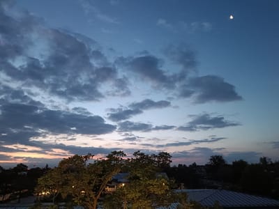 The image depicts a twilight sky over Germering, Germany. The sky is a gradient of deep blue at the top transitioning to lighter shades of blue, pink, and orange near the horizon, indicative of a sunset or sunrise. Dark, textured clouds are scattered across the upper portion of the sky, with a sliver of the moon visible in the upper right quadrant. Below the sky, silhouetted trees with green foliage dominate the foreground and mid-ground. Some of the trees have artificial lights illuminating them from within or below. In the background, the outlines of buildings and other trees are visible, suggesting a suburban or semi-urban environment. The overall scene is tranquil and atmospheric, capturing the soft light of dusk.
