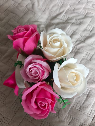 A bouquet of six artificial roses, three pink and three white, is arranged on a textured, light-colored fabric surface, likely a tablecloth or blanket. The bouquet is tied with a white ribbon and a bright pink satin ribbon is visible on the left side. The roses appear to be made of soap or a similar material, and some have small green artificial leaves attached to their stems. The lighting is soft and diffused, casting gentle shadows on the fabric. There are no people or specific landmarks visible in the image, so its location in Sukhodol, Russia cannot be determined.