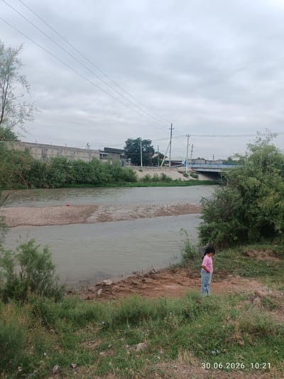 The image shows a wide, murky river flowing through a semi-urban landscape under an overcast sky. On the left bank of the river, there is a gravelly shore with a red object visible in the water. Beyond the river, there are rows of buildings, some appearing to be under construction, and dense green vegetation. A bridge spans the river in the middle ground, with power lines and utility poles visible throughout the scene. On the right bank, a young child wearing a pink shirt and blue jeans stands on the grassy embankment, looking down. The timestamp "30.06.2026 10:21" is superimposed on the bottom right corner of the image. The environment appears to be a somewhat neglected area of a city, with a mix of natural and man-made elements.