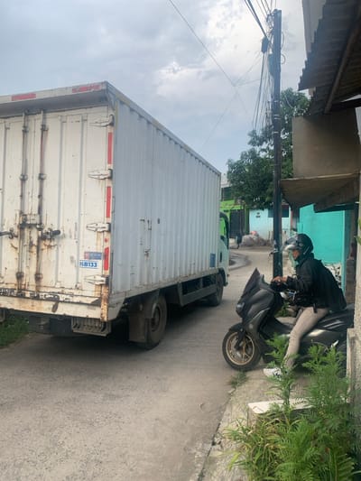 A white delivery truck with the license plate "K8133" is parked on the right side of a narrow street in the city of Lagadar, Indonesia. The truck is facing left and is partially obscured by a building on the right. A person wearing a helmet is sitting on a black scooter facing right and is partially obscured by the truck. The scooter is in the foreground of the image. The person is wearing khaki pants and a black jacket. There are trees on the left side of the street. The street is paved and there is a small amount of green vegetation along the edge of the street. The sky is overcast and there are power lines and a utility pole in the background.