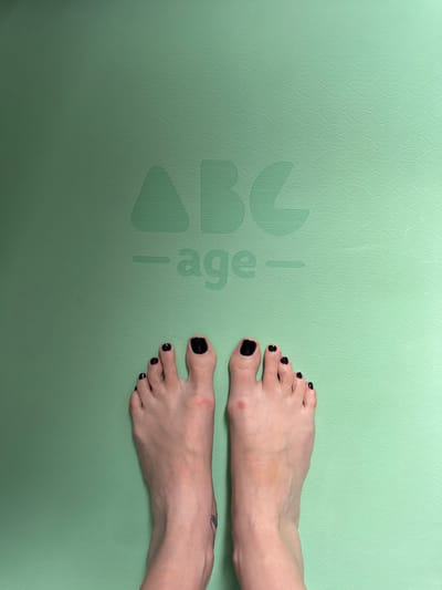 In the image, two bare feet with black nail polish are positioned on a light green textured surface. The surface has a subtle wavy pattern and embossed text that reads "ABC age". The feet are facing upwards, with the toes spread slightly. There are minor blemishes on the skin of the feet, including a red mark on the left foot's big toe and some uneven skin tone on the right foot. A faint tattoo is visible on the left ankle. The overall scene appears to be indoors, possibly on a yoga mat or similar exercise surface. There is no indication of the location being in Боголюбово, Russia.