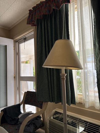 The image shows an upward-angled view of a room interior, likely in a house in Laconia, United States of America. The room has a window with white sheer curtains and green drapes, and a ceiling fan. A lamp with a beige lampshade is positioned in front of the window. Portions of furniture such as chair are visible.