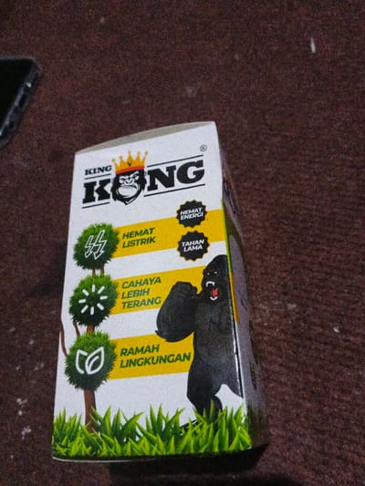 A white box with "KING KONG" written in black letters, with a gorilla wearing a golden crown as the logo. On the side, three green trees with icons representing saving electricity, brighter light, and being environmentally friendly. Next to them, text in Indonesian describes these features. A black gorilla with its mouth open stands powerfully on the box. The bottom of the box is designed with green grass. The box sits on a textured surface with a glimpse of a black phone in the background.