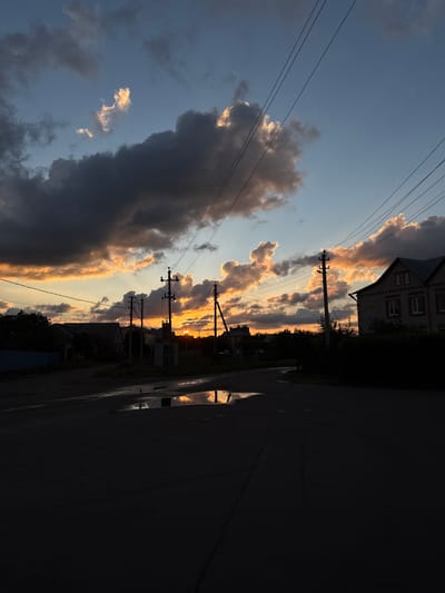 An outdoor, wide-angle, eye-level, vertical shot captures a rural road in Rossosh, Russia, at sunset. The sky is dominated by dark, heavy clouds illuminated by a brilliant orange glow from the setting sun, which is partially visible peeking through the clouds. Thin electrical wires crisscross the frame against the twilight sky. On the right side of the road, a two-story brick house with a gray roof and white-trimmed windows stands in silhouette. Power poles with crossbars and insulators are strategically placed along the road. The asphalt road surface is wet, reflecting the sky and nearby structures in puddles. Trees and other buildings are visible in the middle ground, also silhouetted against the vibrant sky. The overall mood is dramatic and serene, with the natural beauty of the sunset contrasting with the man-made elements of the rural setting.