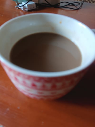 A shallow depth of field, close-up shot focuses on a cup filled with a light brown liquid, likely coffee or tea. The cup has a white interior and a patterned exterior in shades of pink and white. The liquid fills most of the cup, with a slight reflection visible on its surface. The cup rests on a wooden surface, which appears to be a table. In the blurred background, a white electrical plug with prongs is visible, connected to a black cord. The scene is indoors, possibly a home or office environment. There is no indication of the city of Hlobyne, Ukraine, or any specific geographical features in the image.