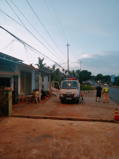The scene is in Xuân Long, Vietnam, at twilight. A white CSGT (Traffic Police) van with flashing red and blue lights is parked partially on a sidewalk. Two men stand on the road; one in civilian clothes, and one in a police uniform. A traffic cone sits nearby. On the sidewalk adjacent to the van, several people are seated around a table in front of a building with a metal roll-up door. Overhead are numerous power lines. The road shows light traffic, including motorcycles.