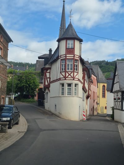 This image depicts a street scene at a corner in the city of Traben-Trarbach, Germany, under a partially cloudy sky. The prominent central feature is a three-story, light-cream colored building with dark red half-timbering, featuring a distinctive corner turret topped with a conical dark slate roof and a weather vane. An inscription "W. P. 19 01" is visible on the turret's second level.

To the left of this central building, an older structure made of rough-hewn reddish-brown stone with darker trim is partially visible, featuring a small round window on its upper section. A black Audi sedan is parked on the paved sidewalk in front of this building. Further down the street to the left, a green street lamp stands near a large, leafy tree, partially obscuring the view of the background. A street sign identifying "Münichstraße" is visible near the tree. Two black waste bins are positioned against a building wall near the corner.

To the right of the central building, a row of structures extends down a winding street. The first building is a pale pink, followed by a yellow building with a tall, narrow window, and then a white building with dark half-timbering, featuring window boxes with red flowers. All visible roofs are dark slate.

The street itself is paved with dark asphalt, with a lighter-colored section indicating recent repair or patch-work. A red and white striped traffic marker stands on the sidewalk near the corner building. Utility lines traverse the upper portion of the frame against the blue sky with scattered white clouds. In the background, rolling hills densely covered with dark green trees rise above the town.