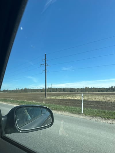 The image depicts a view from inside a vehicle moving on a paved road in a rural area of Jaunolaine, Latvia. The sky is clear and blue with power lines running parallel to the road. A field is visible to the right of the road, with patches of green grass and a line of trees in the distance. A white marker post with a number "3" is located beside the road. The vehicle's side mirror is visible, reflecting the side of the vehicle and the driver's arm. The image is oriented vertically.