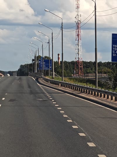 This is a forward-facing, eye-level, medium shot of a highway on a sunny day with scattered clouds. The highway is paved and has white dashed lines indicating lanes. The highway curves to the right in the distance, with a metal guardrail lining the right side. There are several streetlights lining the left side of the highway.

In the background, there is a red and white cell tower, a few trees, and a signpost with directions to various cities, including Torzhok. The signpost appears to be in Russian and lists "ТВЕРЬ", "МОСКВА", "ТОРОПОК", and "С.-ПЕТЕРБУРГ", which correspond to Tver, Moscow, Toropok, and Saint Petersburg, respectively. This indicates the highway is likely near Torzhok, Russia, as it is listed on the directional signage. The environment appears to be rural or semi-rural. There are no people or vehicles clearly visible in the image.