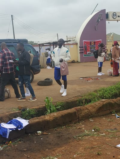The image depicts the exterior of a building with a large, curved, purple archway, likely an entrance to a facility. A banner advertising "NIGERIAN ARMY RECRUITMENT MASSIVE INTAKE" is displayed on the archway. Several individuals, appearing to be recruits or applicants, are present outside. One man in a white jacket and blue shorts walks towards the camera carrying a pink bag. Another man in a black jacket and blue jeans stands near a dark-colored SUV. Other individuals, some carrying backpacks and wearing athletic attire, are visible in the background. The ground is muddy and uneven, with debris and vegetation scattered around. The scene appears to be outdoors, possibly in a public area or near a military installation in Bahit, Nigeria.