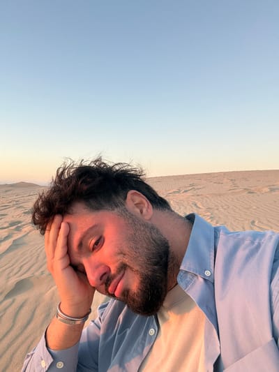 A man is taking a selfie in the sand dunes of La Teste-de-Buch, France. The man has dark, messy hair and a beard, and is wearing a light blue button-down shirt over a beige t-shirt. His left hand is resting on his forehead, and he is looking at the camera with a slight smile. The sand dunes stretch out behind him, with a clear blue sky above. The lighting suggests it is either sunrise or sunset.
