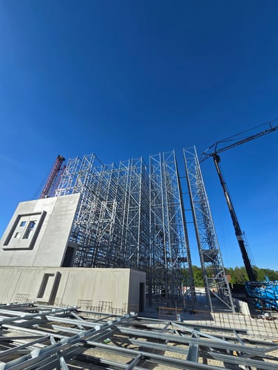 The image displays an active construction site under a clear, vibrant blue sky. The central focus is a massive, intricate steel framework, likely forming the skeleton of a large industrial building or high-bay warehouse. The structure consists of numerous grey metal beams and trusses, indicating multiple levels and extensive internal support.
To the upper left, a prominent black and blue mobile crane extends its long boom across the site. Visible on the crane's boom are the web addresses "www.mammoet.com" and "www.goldbeck.de," identifying Mammoet, a heavy lifting specialist, and Goldbeck, a construction company known for industrial and commercial buildings. A smaller red tower crane is partially visible behind some concrete structures in the lower-left.
In the foreground and right side of the image, partially completed concrete structures and walls are present. Some sections show exposed rebar, indicating ongoing concrete work. One large concrete wall features a distinctive, recessed, stylized 'E' or geometric logo. Another concrete element has a rectangular opening or window frame.
The scene captures a dynamic moment in the construction process, with the steel superstructure well underway and concrete elements simultaneously being formed. The bright sunlight and cloudless sky suggest it is daytime, likely a clear and pleasant day for construction work. The overall impression is of a large-scale building project in progress, consistent with industrial development in Detmold, Germany.