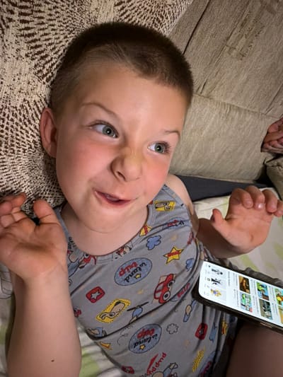 A young boy with short brown hair and green eyes is lying down and looking to his right with a playful expression. He is wearing a grey sleeveless shirt with a pattern of cartoon cars, clouds, stars, and the text "Fun" and "learn" on it. His arms are bent at the elbows, with his left hand raised near his face and his right hand outstretched. A smartphone is resting on his shirt, displaying a grid of images and text, indicating he is likely watching something or browsing on the device. The background consists of a textured, brown couch. The scene appears to be indoors, possibly in a living room. The lighting suggests it is evening or nighttime. The provided context places this scene in Zmeitsa, Bulgaria, though no specific landmarks or city details are visible in the image itself.