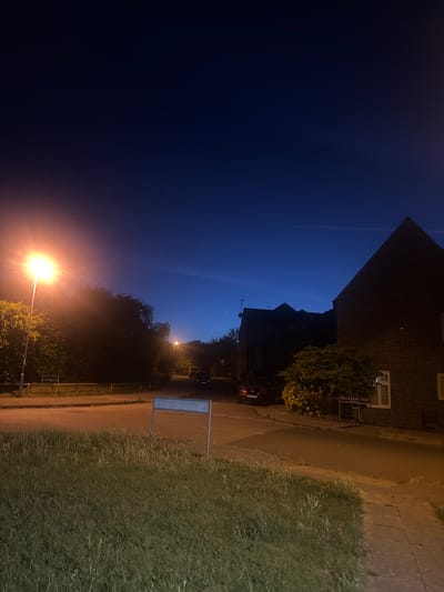 The image depicts a street scene at dusk or night in the Borough of Swale, United Kingdom. A street lamp on the left side of the image illuminates a grassy verge and a paved road. In the background, there are houses, trees, and a few parked cars. A street sign reading "MULBERRY WAY" is visible on the right side of the road. The sky is a deep blue, indicating the transition from day to night.