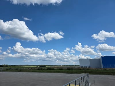 A wide-angle, outdoor, daytime, eye-level shot captures a vast blue sky filled with fluffy white cumulus clouds over a flat, open landscape in Barniewice, Poland. In the foreground, a paved area with light-colored, rectangular stones and white parallel lines is visible. A metal railing with yellow accents runs along the right side of the paved area. Beyond this, a strip of green grass separates the paved area from a distant, flat horizon. The middle ground features a line of trees and scattered buildings, suggesting a rural or semi-urban environment. On the right side of the image, two large, modern industrial buildings are prominent. The building on the left is a light gray structure with a slightly darker gray section and a series of small, dark windows near the base. An external ladder is attached to this building. Adjacent to it, on the right, is a large, vibrant blue building. The overall scene is bright and expansive, dominated by the sky and open space.