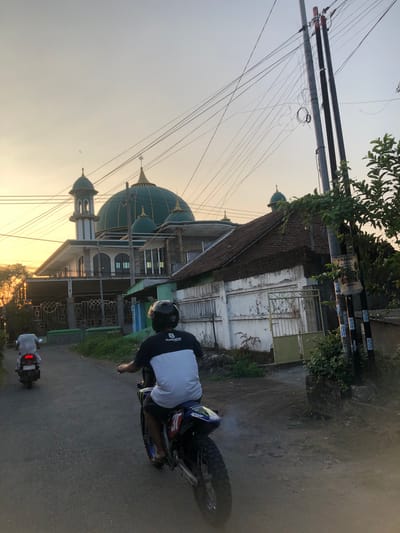 The image shows a street scene in Sentul, Indonesia, during what appears to be late afternoon or early evening, indicated by the warm, low sunlight. A mosque with a large green dome and a tall, slender minaret is prominently visible in the background. In the foreground, two individuals are riding motorcycles on a paved road. The motorcycle closest to the viewer is a dirt bike with a rider wearing a black helmet and a white and black shirt with the Husqvarna logo. The rider is wearing dark shorts. The second motorcycle, further down the road, is a scooter-type bike with a rider wearing a light-colored shirt and dark pants. The environment includes power lines strung across poles, trees, and houses alongside the road.