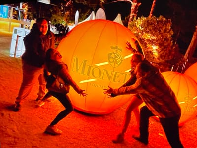 At the "DESIGN DAYS PURKERSDORF" event in Purkersdorf, Austria, four individuals are interacting with a large, illuminated orange inflatable sphere. The sphere displays the text "MIONETTI FONDATA" and a crest with grapes. In the foreground, a person wearing dark leggings and sneakers leans back with their arms outstretched towards the sphere, appearing to embrace it. To their right, another individual in a patterned jacket also has their arms outstretched, seemingly touching the sphere. Behind them, a man in a dark hoodie and light-colored pants is visible, smiling and gesturing towards the sphere. Another person, partially obscured, stands to the left, also facing the sphere. The ground is covered in a reddish-orange material, possibly gravel or dirt, and illuminated by the vibrant light of the sphere. In the background, trees with orange lights and white tents suggest an outdoor nighttime event.