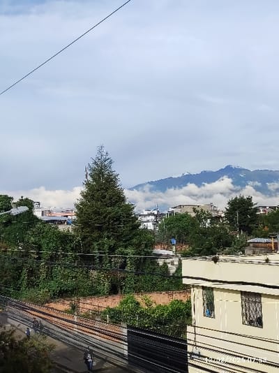 This image presents an elevated, wide-angle view of an urban or suburban area within Puembo, Ecuador, captured on 12/02/2026 at 07:58.

The upper portion of the frame consists of a light blue sky with significant white and grey cloud cover. A single, dark, thin cable diagonally traverses the sky from the upper-left to the mid-right.

In the background, a distant mountain range is visible, its peaks partially obscured by thick white clouds. Lighter areas on the higher slopes suggest snow or exposed rock.

The midground is characterized by a dense arrangement of buildings, primarily light-colored, with various rooflines. These structures are interspersed with abundant green foliage, including numerous trees and bushes. A prominent, tall, dark green conifer stands centrally within this vegetated area. A network of black overhead power lines and utility poles is visible, crisscrossing horizontally and diagonally across the midground. Below the main cluster of trees and buildings, a horizontal reddish-brown brick wall or fence is present.

The foreground depicts a paved area or street in the lower-left, featuring multiple dark, parallel lines resembling tracks or heavy-duty ground cables. Three individuals are visible on this surface: one person in a light-colored top and dark trousers walks left-ward, a second person is partially obscured further left, and a third person in dark clothing walks right-ward. The lower-right foreground shows a section of a cream-colored building with dark window frames and metal security grates. The flat roof of this building contains dark, unidentifiable debris or organic matter. Several additional black utility lines run horizontally across the front of this building.