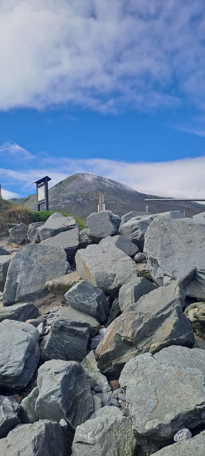 The image shows a rocky shoreline with a mountain in the background. In the foreground, there are large, grey rocks scattered on the sand. A wooden signpost with information is visible to the left of the mountain. The sky is blue with scattered white clouds. The mountain is covered in grass and has some fog around its peak. The location is Pier Road, Ireland.