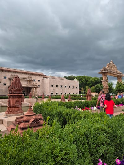 This image, taken in Robbinsville Township, United States of America, captures a scene outside a large, light-colored building constructed with stone. The building has multiple rows of square windows and is adorned with decorative architectural elements. In the foreground, a man and a child are walking through a manicured garden area. The man is wearing a red and white plaid shirt and khaki pants, while the child is wearing a bright red shirt and blue jeans. They are walking towards the right side of the frame, away from the camera, passing by meticulously trimmed green bushes and colorful flowers. To the right of the couple, a large, ornate stone archway stands, decorated with intricate carvings. Several other decorative red stone structures, resembling small temples or stupas, are visible throughout the garden, some are located near a water feature to the left of the main building. The sky is overcast with dark, dramatic clouds, suggesting a potentially stormy day. Other people can be seen in the background, suggesting the location is a public space. The overall scene depicts a serene, yet imposing, architectural and garden setting under a moody sky.