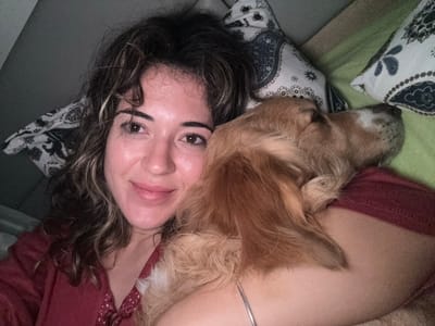 An adult female and a golden-colored dog are depicted in a close-up indoor scene within the city of Kalyvia Thorikou Municipal Unit, Greece. The female, positioned to the left of the frame, has medium-length wavy brown hair with lighter highlights, brown eyes, and is looking directly at the camera with a slight smile. She wears a maroon long-sleeved garment, with a subtle pattern visible near the neckline. A silver-colored bracelet is present on her left wrist. The dog, with golden-brown fur and a lighter, possibly white, snout area, is resting its head against the female's left shoulder and chest, appearing to be asleep or highly relaxed. The background consists of soft furnishings, including a white pillow with a dark, intricate pattern on the left, and a partial view of a green pillow or blanket on the right. The overall lighting is dim, indicating an evening or low-light indoor environment.