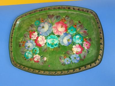A vintage, oval, green metal tray with a decorative floral painting is displayed against a blue background, with a sliver of yellow visible in the upper left corner. The tray features a detailed border painted with gold leaf motifs. The central painted design consists of a cluster of colorful flowers in shades of pink, red, blue, and teal, interspersed with green leaves and small orange berries. The tray appears to be worn, with some scratches and fading visible on its surface. The scene is devoid of people or any indication of human activity. There is no information in the image that suggests a connection to Zhirnovsk, Russia.