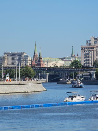 The image shows a body of water with several boats on it, as well as a cityscape in the background. The water appears to be a river, and there is a long blue floating barrier in the foreground. On the water, there are two white motorboats, each with an awning and several people on board. In the distance, a larger white and blue passenger boat is visible, with more people on its decks.
The cityscape includes several buildings, including what appear to be historical structures with green spires and red brick walls, likely part of the Kremlin. A bridge with metal supports spans the water in front of these buildings. The sky is clear and blue, indicating a sunny day. On the bank in the middle ground, there are trees, lampposts, and several people, some of whom are wearing orange vests, suggesting construction or maintenance work. The overall scene is one of a bustling urban environment on a clear day, with a focus on the water and the boats. The location is identified as Балтия Кп, Russia.