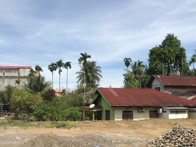 This image captures a daytime scene in Lhoksukon, Indonesia, depicting a semi-urban or rural landscape under a partly cloudy sky. The foreground consists of uneven, bare, light brown earth, suggesting an undeveloped or recently disturbed area, with a pile of light-colored rocks on the right.
In the midground, several houses are nestled among dense tropical vegetation. A prominent single-story house in the center-right features light green walls and a rusty reddish-brown corrugated metal roof. This house has louvered windows, an outdoor air conditioning unit, and a white satellite dish mounted on a smaller, attached porch area. Another building with a similar reddish-brown roof is partially visible behind it to the right. On the far left, a light-colored concrete structure with a red roof is partially seen, possibly unfinished or under construction.
The scene is abundant with greenery, dominated by numerous tall, slender palm trees (likely areca palms) and broader-leaved coconut palms, interspersed with various bushes and other trees. The sky is a light blue with scattered white clouds, indicating bright daylight. In the distant background on the left, hints of a structure with blue, dome-like roofs are visible, characteristic of a mosque, further reinforcing the geographical context. No people are present, and there is no explicit activity taking place. No visible text is discernible in the image.