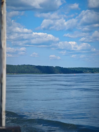 The image depicts a wide, calm river on a sunny day with scattered white clouds in a blue sky. The left edge of the frame is partially obscured by a vertical, weathered wooden structure, possibly part of a pier or railing. The river water exhibits gentle ripples and reflects the sky and surrounding landscape. Across the river, a dense, dark green forest stretches along the horizon, meeting the water's edge. A small, orange buoy is visible in the water near the center of the frame. In the distance, a few houses with red roofs are nestled within the treeline. The scene appears to be a serene natural landscape. The location is the city of Krutoy Log, Russia.