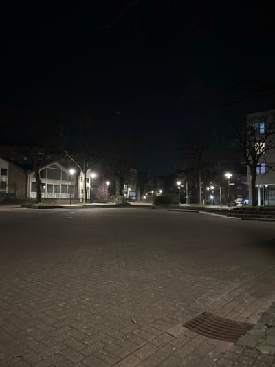 This image depicts a nighttime view of a public square in the Dahl district of Hagen, Germany, specifically located in front of the Jugendzentrum Dahl at Dahl Buschmühle 3, 58091 Hagen. The scene is illuminated by artificial light sources under a dark, clear sky, where a single prominent star-like object is visible near the upper center-left.

The foreground and midground consist of a large, empty square paved with reddish-brown interlocking pavers, exhibiting minor wear and scattered debris. A rectangular metal storm drain grate is visible in the lower right corner of the paved area.

On the left side of the square, a two-story building features a light-colored facade and a distinctive A-frame roof section with a large, multi-paned window; this is the Jugendzentrum Dahl. Its windows appear dimly lit or reflective. Bare deciduous trees, indicating a dormant season, are distributed across the square and along its edges, with their dark branches silhouetted against the ambient light.

On the right side, a multi-story, light-colored modern building stands with several rectangular windows, some of which are illuminated with blue or white interior light. Numerous pole-mounted streetlights with white-yellow light sources are positioned throughout the square and along the distant street, casting light and shadows across the environment. Other less distinct buildings and structures are visible further down the illuminated street in the background. No people or vehicles are present in the scene.