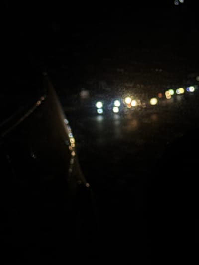A dark, blurry image with out-of-focus lights resembling street lamps or city lights in the distance. A diagonal line, also out of focus and possibly a reflection, cuts across the lower part of the image. The overall scene is obscured by darkness and lack of clarity, making it difficult to discern specific details. It's likely a nighttime scene viewed through a window or a similar obscuring medium.