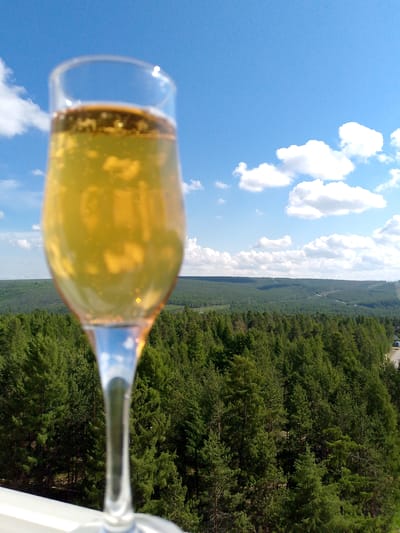 The image shows a glass of champagne in the foreground, with a lush green forest and a bright blue sky with white clouds in the background. The champagne is a golden color with small bubbles rising to the surface. The forest is dense and appears to be a coniferous forest. The sky is clear and sunny, with a few fluffy white clouds scattered across it. The overall impression is one of a peaceful and celebratory scene in nature. This scene could be located in the city of Neryungri, Russia, which is known for its vast forests and clear skies.