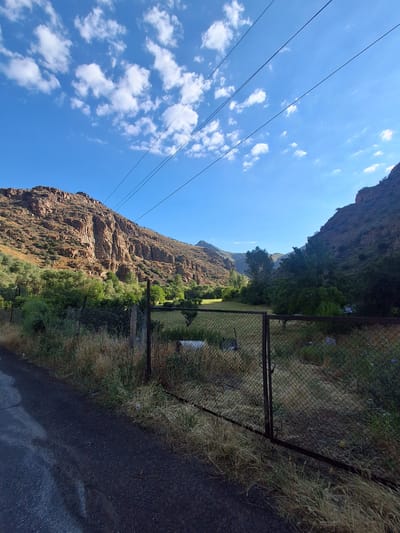 The image depicts a rural landscape with a road in the foreground, leading towards a fenced area. Beyond the fence, a grassy field is visible, bordered by trees and the foothills of rugged, rocky mountains. Power lines stretch across the bright blue sky, which is dotted with white, fluffy clouds. The overall impression is one of a serene, sun-drenched day in a mountainous region, likely Vayk, Armenia, given the characteristic landscape. There are no people or animals in the frame, and the scene appears to be undisturbed.
