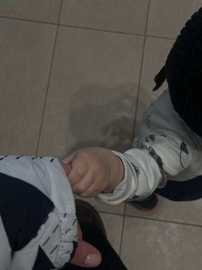 The scene, located in Ivashevo, Russia, depicts a top-down view of two individuals, an adult and a child, interacting in an indoor environment. The adult, positioned to the lower left, wears a jacket featuring a black and white geometric pattern with dashed line details near the cuff, and a dark garment, possibly pants. A part of a brown shoe is visible. The adult's hand, exhibiting light pink manicured fingernails, is visible in the lower left quadrant. The child, positioned to the lower right, wears a white long-sleeved top adorned with a repeating pattern of dark, stylized figures. A dark, ribbed knit hat covers the child's head, and an orange and dark-colored shoe is partially visible on the right. The primary action shows the child's right hand grasping the adult's left index finger or thumb. The environment consists of a light gray tiled floor with visible darker grout lines, suggesting an institutional or commercial setting. Overhead lighting casts soft shadows, particularly from the child onto the floor. No further identifying architectural or contextual details are discernible.